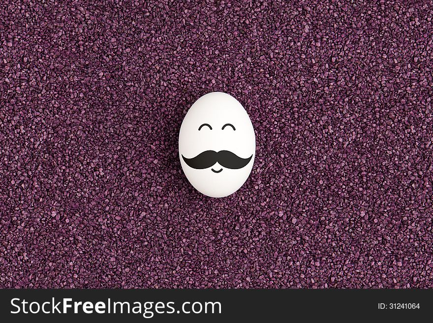 A single egg with mustache are lying on the decorative purple sand and smiling. A single egg with mustache are lying on the decorative purple sand and smiling.