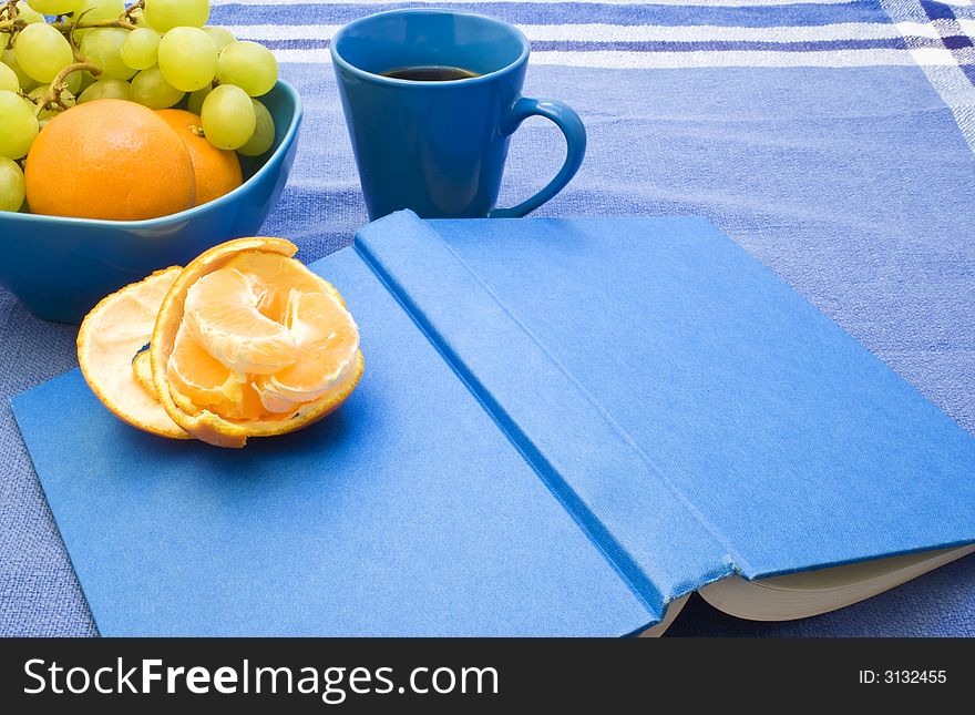A book and a cup of coffee and some fruit on a blue tablecloth. A book and a cup of coffee and some fruit on a blue tablecloth.