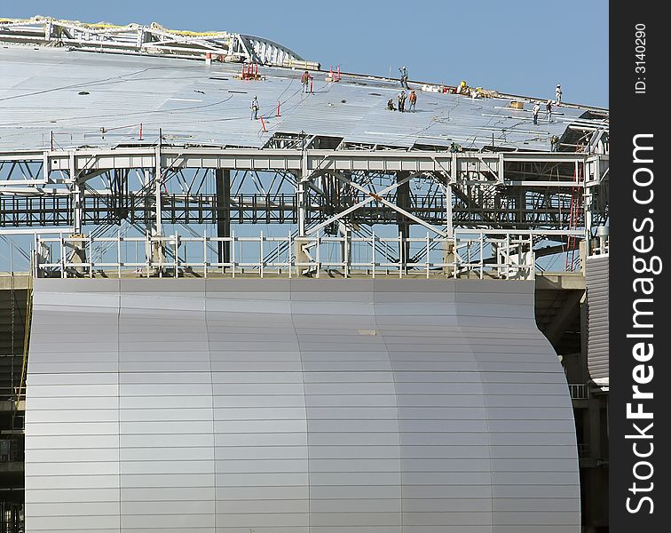 People dwarfed by the roof and side of a stadium under construction - people on root - blue sky background. People dwarfed by the roof and side of a stadium under construction - people on root - blue sky background.