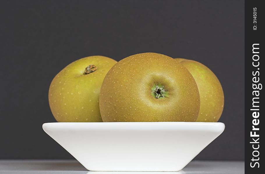 A white bowl containing three golden Russet apples, against a dark background. A white bowl containing three golden Russet apples, against a dark background.