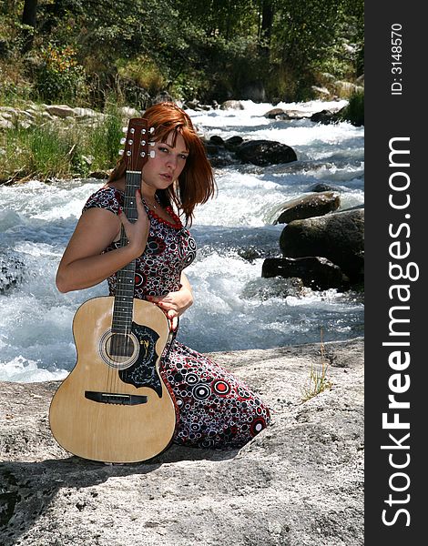 The girl with a guitar sits on a stone on coast of the mountain river in clear day