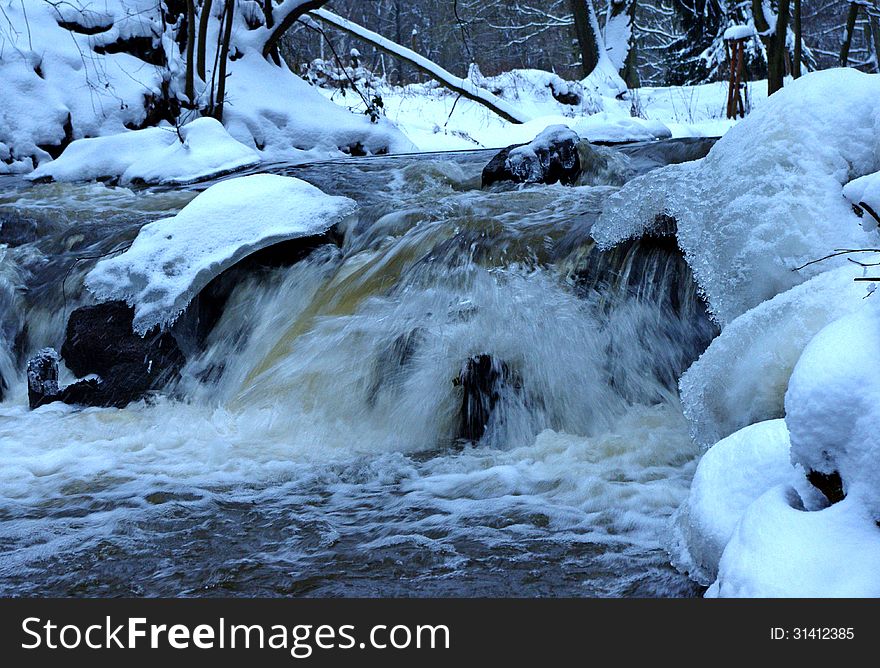 The photograph shows a small waterfall. It is winter on the banks and in the surrounding trees behind a layer of snow. The photograph shows a small waterfall. It is winter on the banks and in the surrounding trees behind a layer of snow.