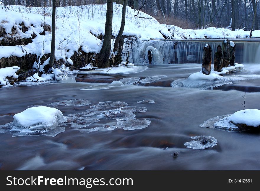 The photograph shows a small waterfall. It is winter on the banks and in the surrounding trees behind a layer of snow. The photograph shows a small waterfall. It is winter on the banks and in the surrounding trees behind a layer of snow.