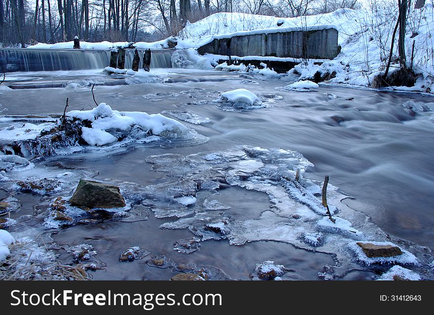 The photograph shows a small waterfall. It is winter on the banks and in the surrounding trees behind a layer of snow. The photograph shows a small waterfall. It is winter on the banks and in the surrounding trees behind a layer of snow.
