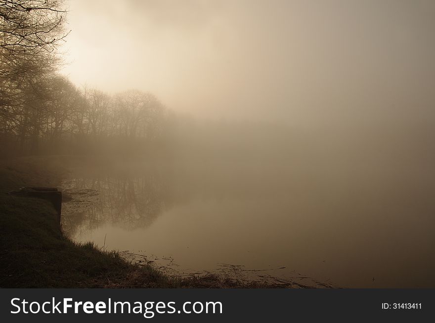 The photograph shows a foggy morning. Above the ground rises a dense layer of fog lit up by the rising sun. In the fog you can see hazy outlines of leafless, naked trees.