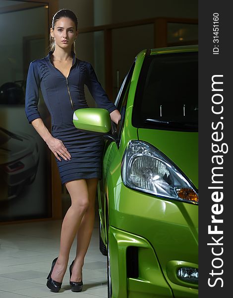 Good looking female model wearing fashionable dress and high heel shoes standing near by new and modern, bright green color, automobile. Indoors, auto salon background. Good looking female model wearing fashionable dress and high heel shoes standing near by new and modern, bright green color, automobile. Indoors, auto salon background.