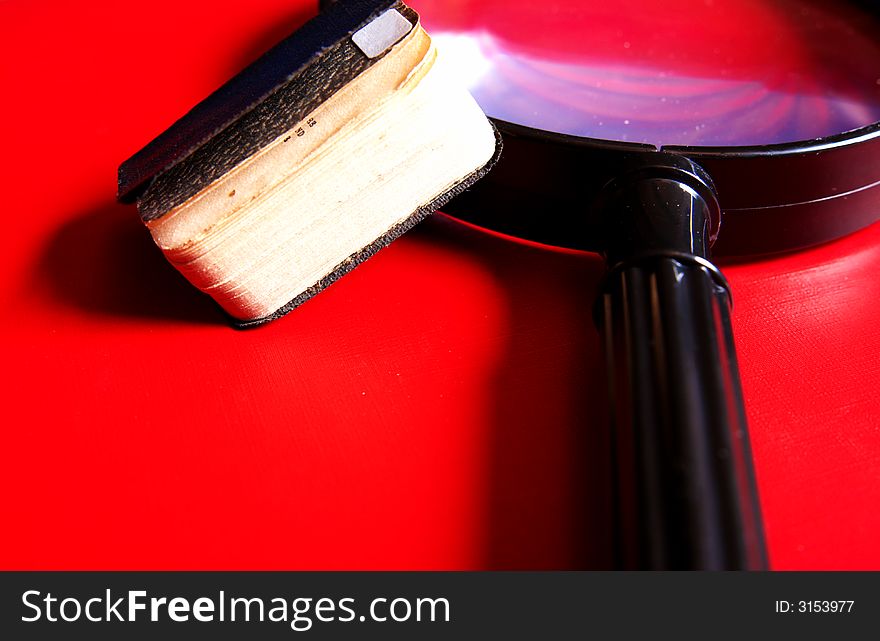 Small black book lying on a big magnifying glass on a red background. Small black book lying on a big magnifying glass on a red background
