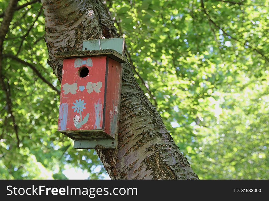 Red birdhouse with painted decorations hangs from tree trunk with green foliage in the background. Red birdhouse with painted decorations hangs from tree trunk with green foliage in the background.