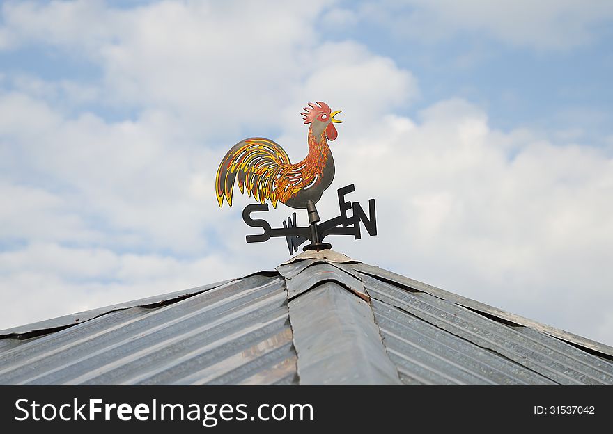 Colorful weather vane with cock figure on the roof over cloudy sky