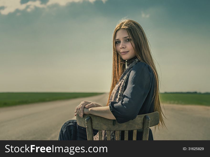 Beautiful Girl With Long Hair Sits Outdoors On A Chair On The Road