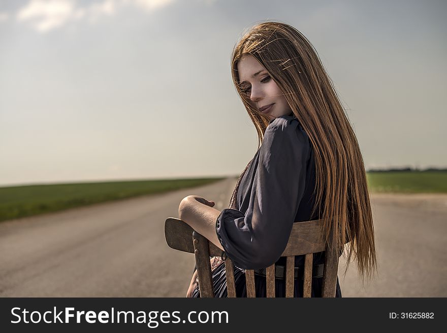 Beautiful Girl With Long Hair Sits Outdoors On A Chair On The Road