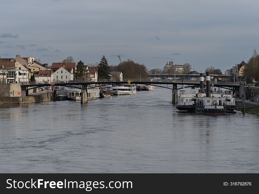 In the frame there is a pedestrian bridge over the Danube River in the city of Regensburg, Germany.  There is a passenger pier nearby.  There are several river-class passenger ships at the berth.