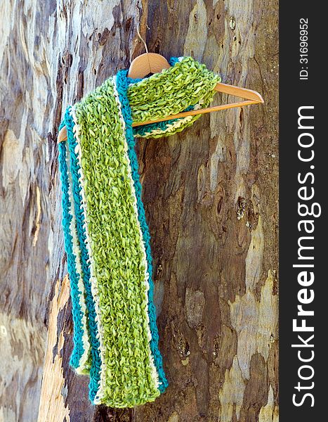 Bright green and blue crochet scarf, fluffy and warm hanging on hanger against wooden tea tree background; concept of winter is coming