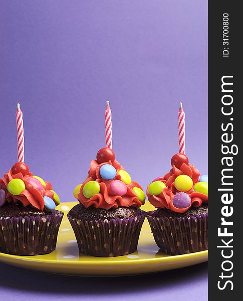 Three bright candy covered cupcakes with birthday candles on yellow polka dot plate against a purple background for Childrens Birthday, Halloween trick or treat, or Christmas party. Vertical with copy space for your text here. Three bright candy covered cupcakes with birthday candles on yellow polka dot plate against a purple background for Childrens Birthday, Halloween trick or treat, or Christmas party. Vertical with copy space for your text here.