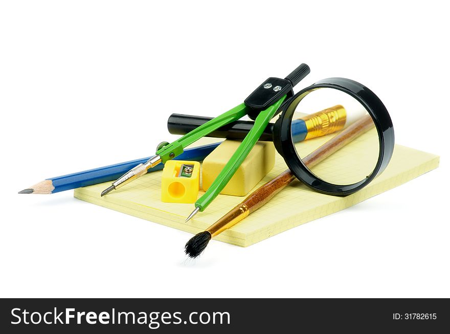 Arrangement of Pencil, Pen, Paintbrush, Pencil Sharpener, Eraser, Drawing Compass and Magnifier on Yellow Note Pad isolated on white background. Arrangement of Pencil, Pen, Paintbrush, Pencil Sharpener, Eraser, Drawing Compass and Magnifier on Yellow Note Pad isolated on white background