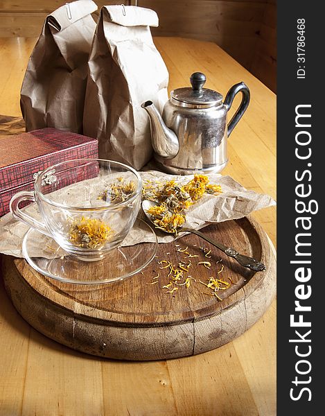 A glass cup, teespoon, kettle and dried marigold flowers on a wooden board. A glass cup, teespoon, kettle and dried marigold flowers on a wooden board.