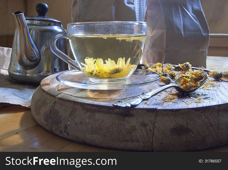 A cup of herbal tea. Near kettle, tea spoon and dried marigold flowers on a wooden board.