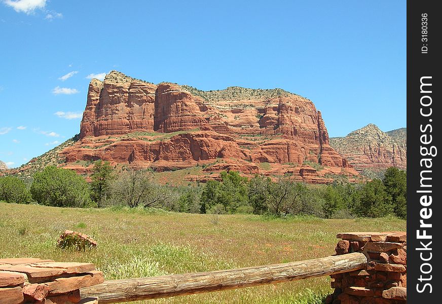 Red Rock formation in the background against a deep blue sky. Trees and field in the midground and a rustic fence in the foreground. Red Rock formation in the background against a deep blue sky. Trees and field in the midground and a rustic fence in the foreground.