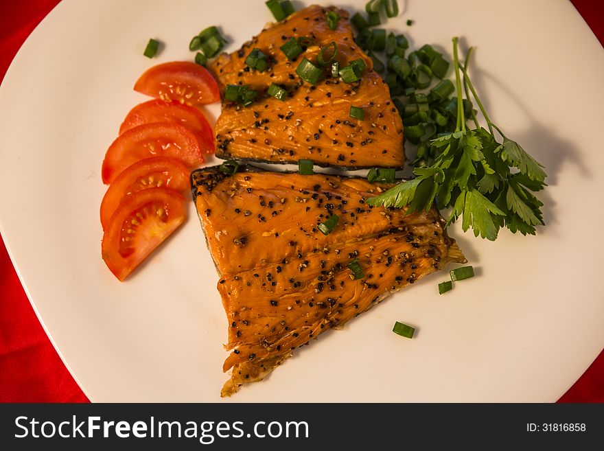 Delicious smoked fish with spices on top served with juicy tomato slices, parsley and green onions. White plate background. Delicious smoked fish with spices on top served with juicy tomato slices, parsley and green onions. White plate background.
