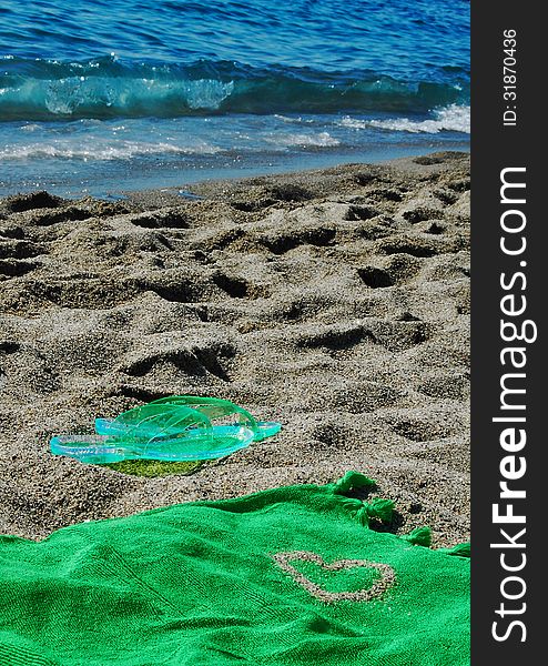 Green flipflop on a beach covered by sand, towel with a heart made by sand and the sea in the background. Green flipflop on a beach covered by sand, towel with a heart made by sand and the sea in the background