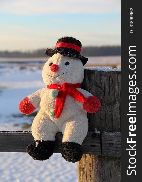 Toy snowman sitting on a wooden fence outdoors at sunset in winter. Toy snowman sitting on a wooden fence outdoors at sunset in winter