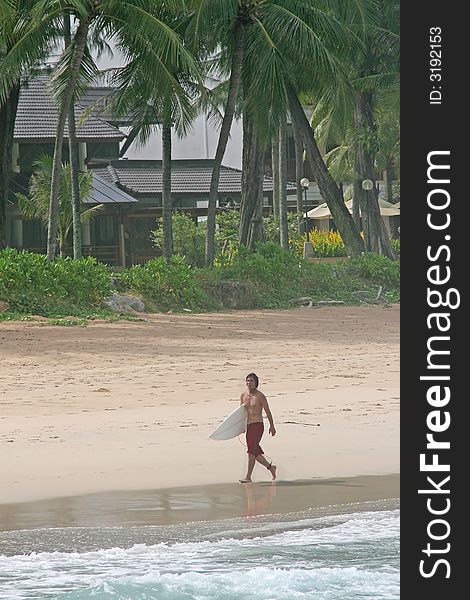 A surfer walks along the waters edge on a tropical beach in Thailand. A surfer walks along the waters edge on a tropical beach in Thailand