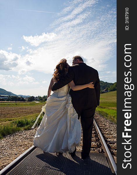 Bridal Couple (over 40 years old) amorous from the back. They are hugging each other while standing on a railway track. It is summer, great sky. Bridal Couple (over 40 years old) amorous from the back. They are hugging each other while standing on a railway track. It is summer, great sky.