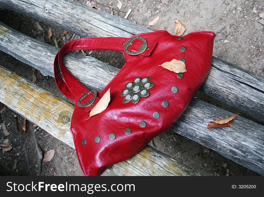 Red leather bag and dry leaf on a wooden bench in the forest