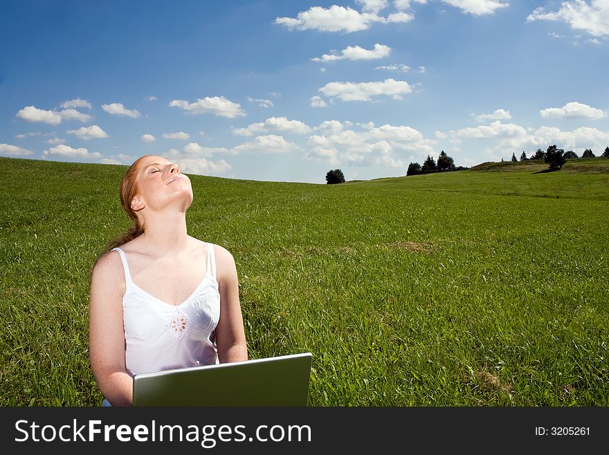 Woman sitting with laptop on a meadow going to the horizon. Blue sky with white clouds. Face towards the sun, eyes closed. Woman sitting with laptop on a meadow going to the horizon. Blue sky with white clouds. Face towards the sun, eyes closed.