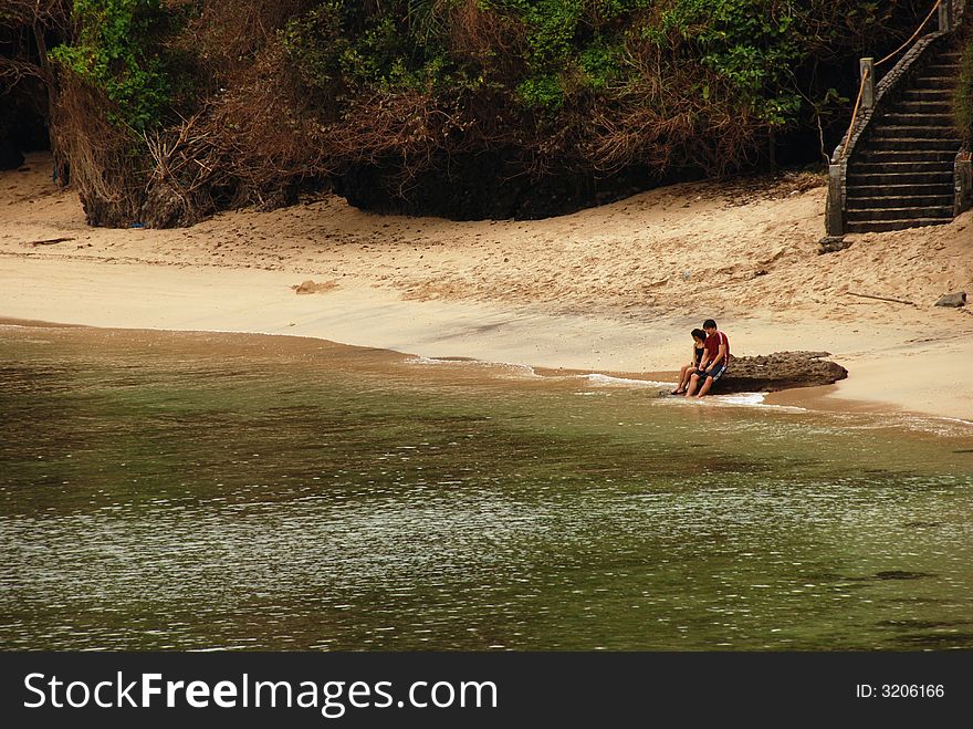 Young couple sitting by a rock with their feet on the water on the beach. Young couple sitting by a rock with their feet on the water on the beach