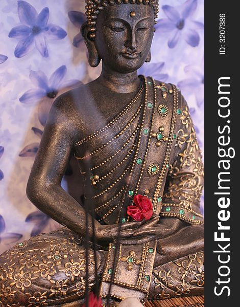 Meditating buddha statue with lilac background holding a red flower and burning incense sticks in front. Meditating buddha statue with lilac background holding a red flower and burning incense sticks in front