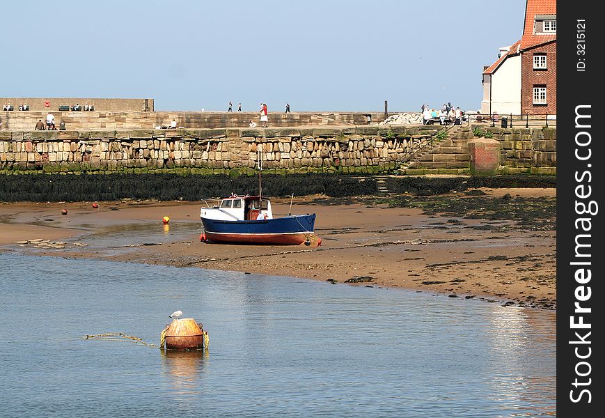 Whitby harbor view showing beached fishing boat. Tourists walk along sea wall in background. Whitby harbor view showing beached fishing boat. Tourists walk along sea wall in background.