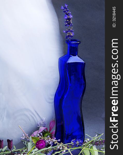 Blue curvy bottle with fresh flowers at the base with a wind blown white curtain fluttering. Blue curvy bottle with fresh flowers at the base with a wind blown white curtain fluttering