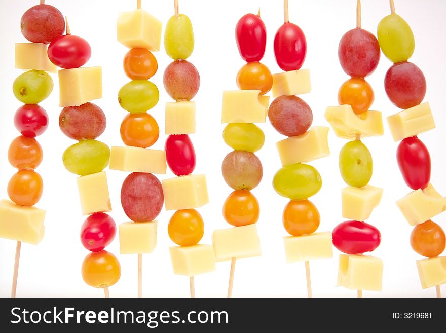 A row of fruit shashliks made with grapes, physalis, tomatoes and cheese, on a white background.