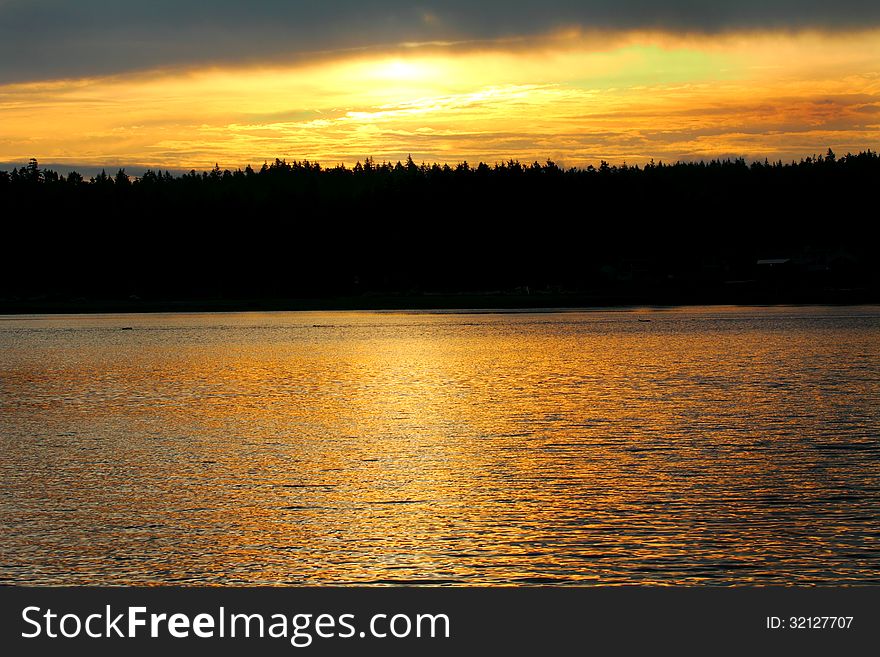 A golden sunset reflection over a large body of water, silhouette of forest in background. A golden sunset reflection over a large body of water, silhouette of forest in background.