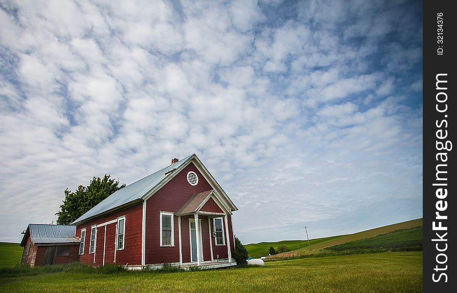 Little red schoolhouse on green grass with blue clouded sky in background.