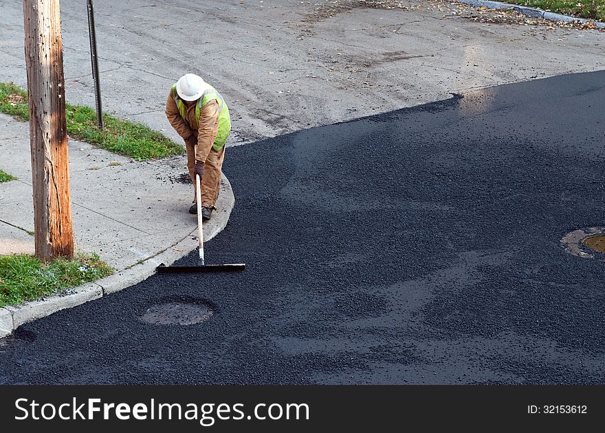 Construction worker putting the finishing touches on repaving a street by manually spreading the asphalt on top.