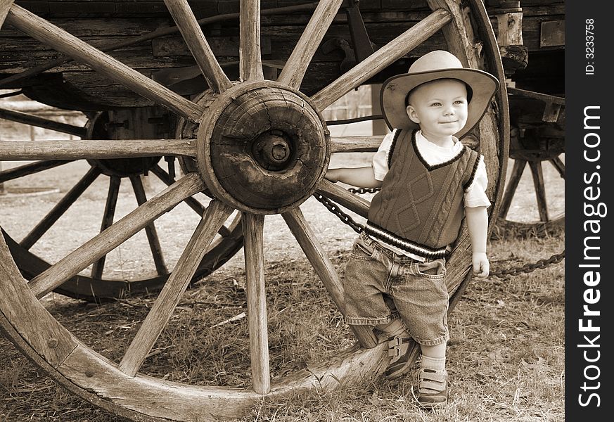 A baby boy hangs out by a large wheel on an old wagon on the farm with cowboyhat and all. A baby boy hangs out by a large wheel on an old wagon on the farm with cowboyhat and all.