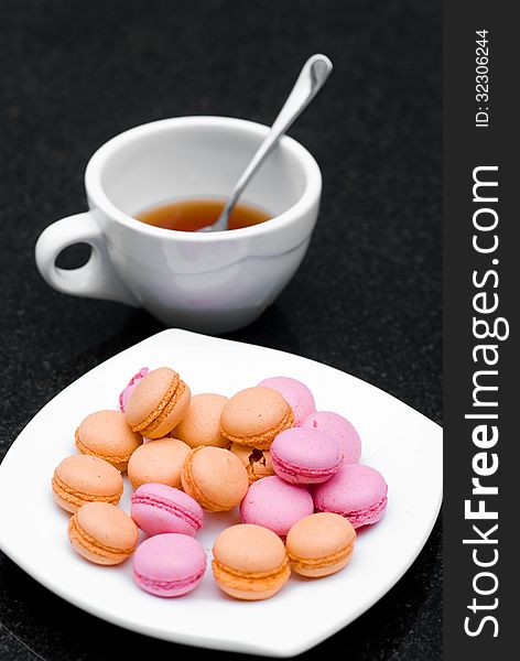 Pile of pink and orange macaroon in a plate with a cup of tea on black table. Pile of pink and orange macaroon in a plate with a cup of tea on black table