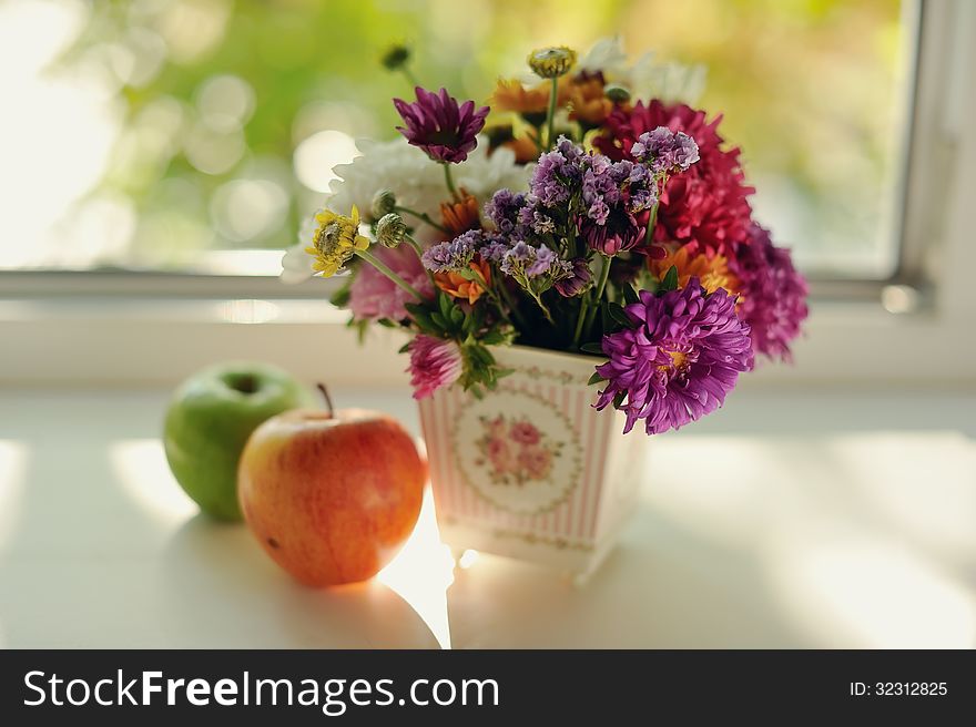 On the window sill is a bouquet of wild flowers in a beautiful decorative vase and some apples are. On the window sill is a bouquet of wild flowers in a beautiful decorative vase and some apples are