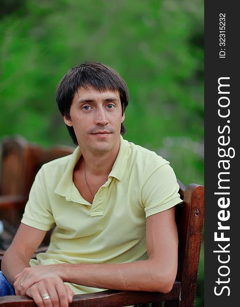 A young man in a yellow shirt is sitting on a wooden chair in nature. A young man in a yellow shirt is sitting on a wooden chair in nature