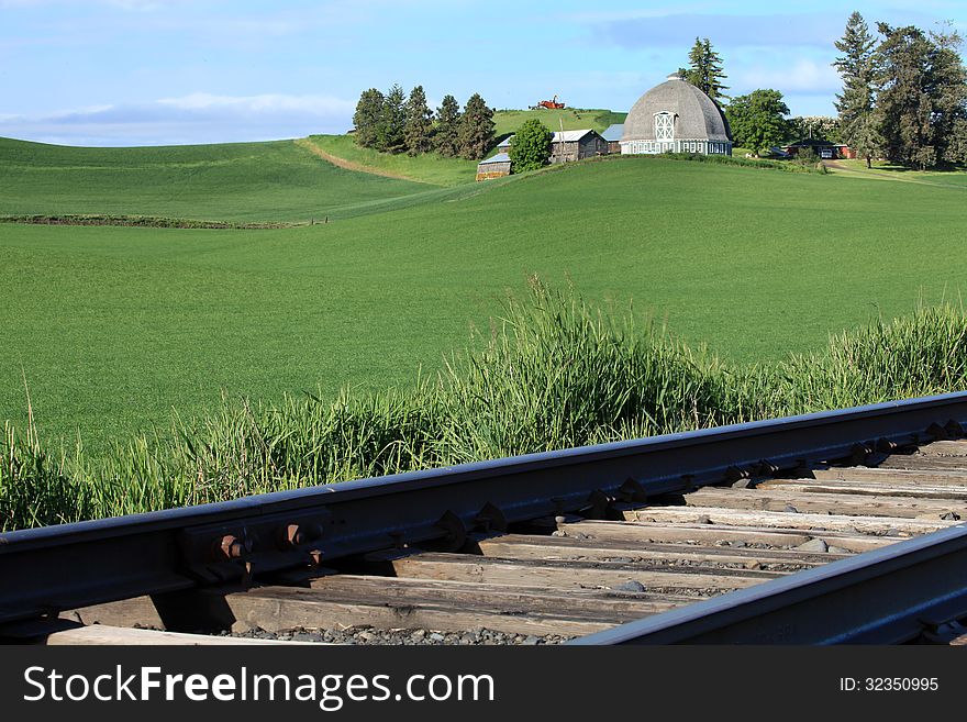 View from Palouse area of Washington State with railroad track in foreground and farmstead surrounded by green field in background. View from Palouse area of Washington State with railroad track in foreground and farmstead surrounded by green field in background.