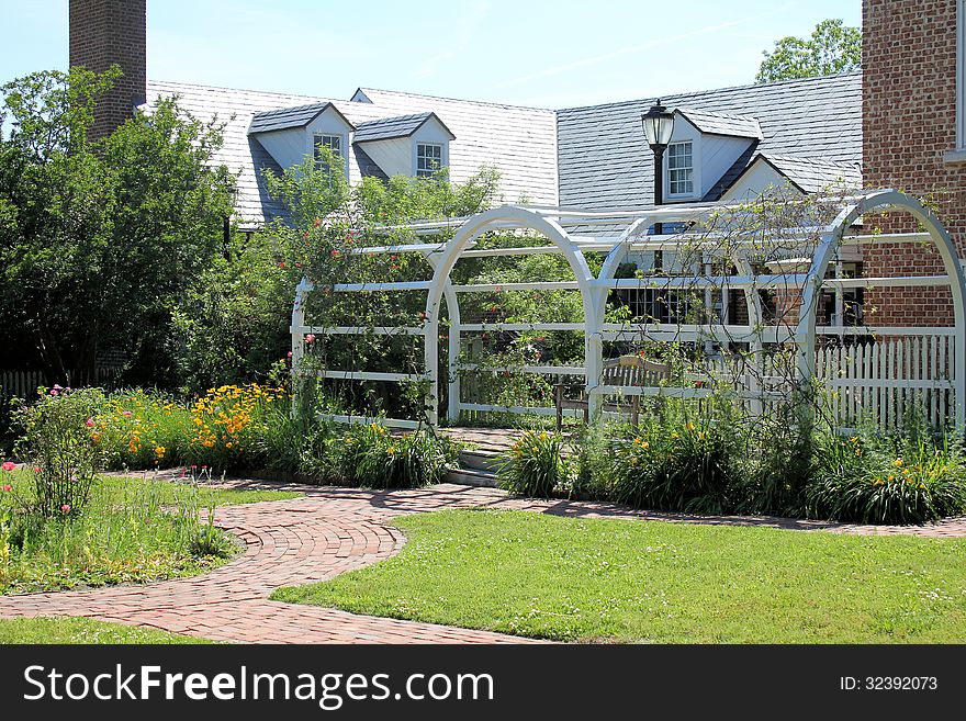 White arbor in garden with brick building in background and brick walkway in foreground.