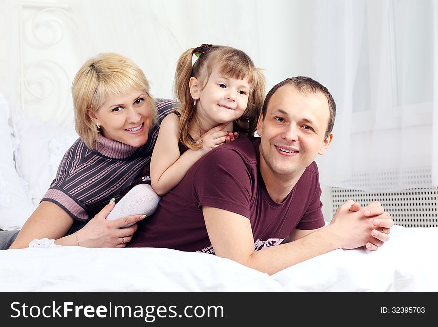 Little girl, father and mother lie on white double bed and smile. Focus on woman. Little girl, father and mother lie on white double bed and smile. Focus on woman.