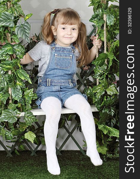 Happy little girl in denim jumpsuit sits on swing under green ivy.