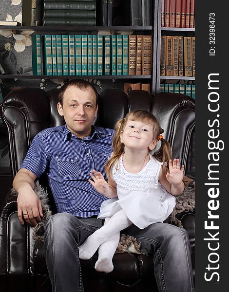 Man and her daughter sit in leather armchair next to shelves with lots of books.