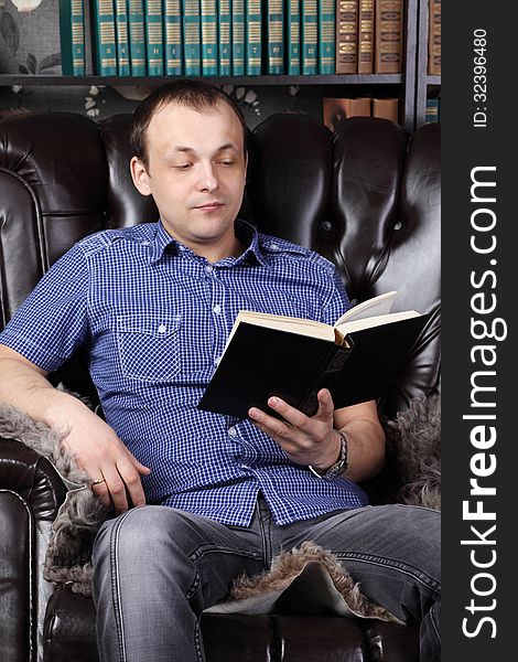 Young man sits in leather armchair and reads book next to shelves with lots of books.