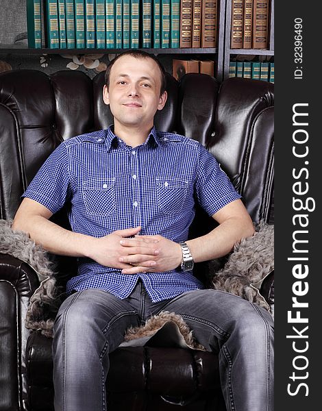 Young man sits in leather armchair next to shelves with lots of books.