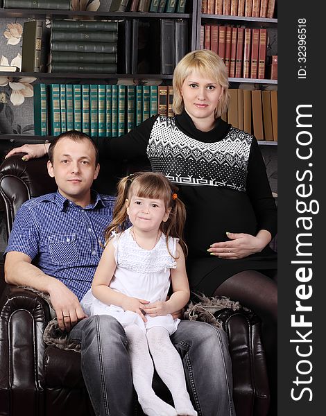 Father, mother and daughter sit in leather armchair next to shelves with books. Father, mother and daughter sit in leather armchair next to shelves with books.