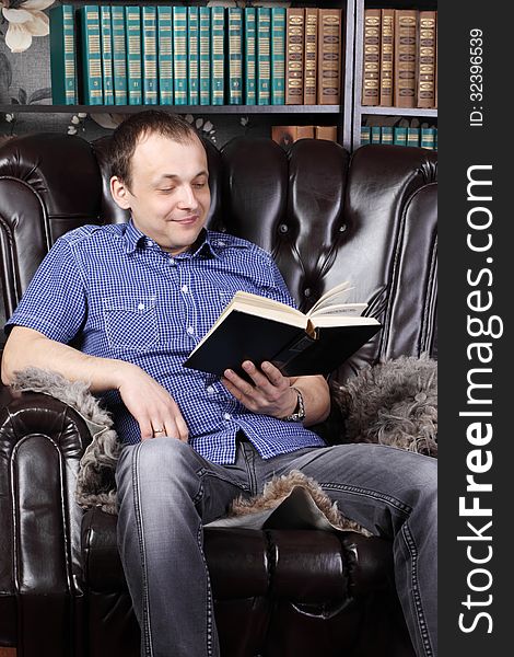 Smiling man sits in leather armchair and reads book next to shelves with lots of books.